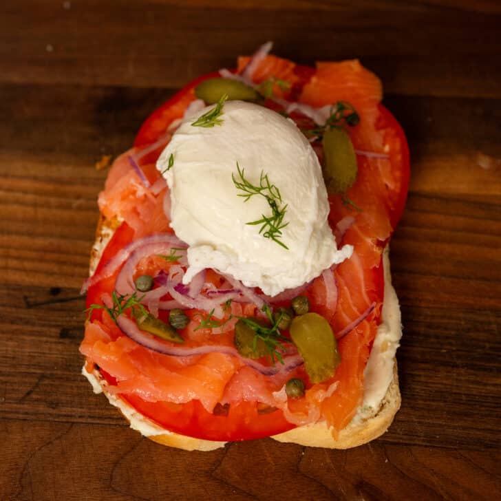 Piece of sourdough toast topped with tomato, smoked salmon, poached egg, red onion, dill, capers and cornichons